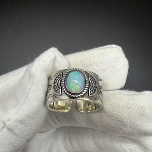 May include: A wide silver ring featuring a central oval-shaped iridescent opal cabochon surrounded by a twisted rope border. The band is decorated with etched geometric patterns and semi-circular motifs on both sides of the stone setting.