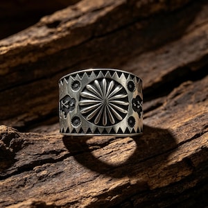 May include: A wide silver ring featuring a central sunburst motif surrounded by small circular stamps and geometric triangular borders. The metal surface has a darkened, oxidized finish that highlights the intricate engraved detailing.
