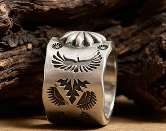 Könnte beinhalten: Ein silberner Ring mit detaillierten, von amerikanischen Ureinwohnern inspirierten Designs. Der Ring zeigt ein zentrales Vogelmotiv mit ausgebreiteten Flügeln, florale Akzente und ein strukturiertes Band. Der Ring wird vor einem natürlichen Holzhintergrund präsentiert.