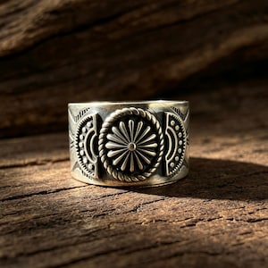 May include: A wide silver ring featuring a central circular medallion with a sunburst pattern surrounded by a twisted rope border. The band is decorated with symmetrical semi-circular motifs and small engraved dots on both sides of the center.