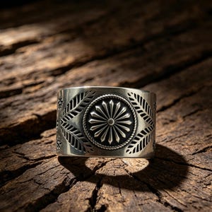 Op de afbeelding: Een brede zilveren ring met een centraal rond medaillon met een zonnestraalpatroon, omringd door een gedraaide touwrand. De band is versierd met gegraveerde bladmotieven aan beide zijden van het medaillon en kleine sterachtige accenten.