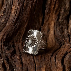Peut inclure: Une bague en argent avec une face rectangulaire ornée d'un motif de soleil. La bague présente des motifs détaillés sur les bords et sur le corps. La bague est présentée sur une surface en bois foncé texturée.