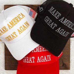 May include: Three baseball caps in white, black, and red, each embroidered with "MAKE AMERICA GREAT AGAIN." The white cap has gold lettering, the black cap has gray lettering, and the red cap has white lettering. Each cap features an American flag.