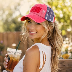 May include: A red baseball cap featuring a distressed finish, a rectangular American flag patch on the front, and a blue mesh back panel adorned with white stars. The cap has a curved brim and a structured crown with visible stitching details.
