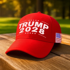 May include: A red baseball cap with the text "TRUMP 2028" in white, along with a small American flag patch. The cap is sitting on a wooden surface, with a blurred green background.