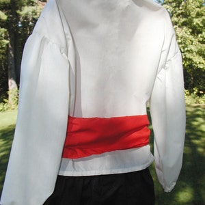 May include: White long-sleeved shirt with a red sash around the waist. The shirt has a gathered puff sleeve detail.