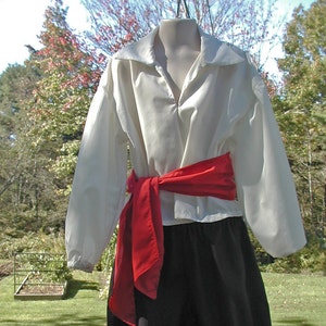 May include: A white shirt with a red sash tied around the waist. The shirt has a collar and long sleeves. The sash is tied in a bow at the front.