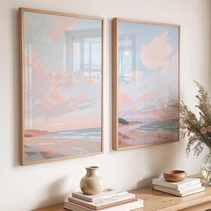 May include: Two framed landscape art prints depicting a beach scene with pastel colors of pink, blue, and peach. The artwork is displayed on a white wall above a wooden table with decorative items.