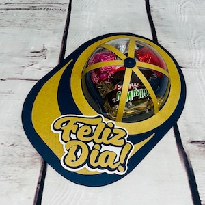 May include: A gift box with a navy blue and gold design, shaped like a baseball cap. The box contains wrapped chocolates and the words "Feliz Dia!" in gold script.