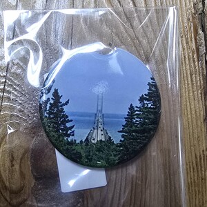 May include: A round button featuring a scenic image of a bridge over water, framed by green trees. The button is encased in clear plastic, suggesting it's a collectible or souvenir item. The background is a wooden surface.