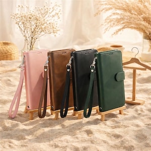 Puede incluir: Cuatro fundas de teléfono de cuero en rosa, marrón, negro y verde, cada una con una correa de muñeca y un cierre de botón a presión. Las fundas se exhiben en pequeños caballetes de madera, sobre un fondo arenoso con plantas decorativas.