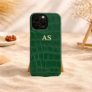 May include: A green phone case with a crocodile skin texture and the gold letters "AS". The case has a camera cutout and is displayed on a sandy surface. The case is propped up on a wooden stand.