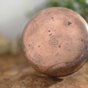 Puede incluir: La parte inferior de una olla de cobre con una base circular ligeramente abollada. La olla tiene una pequeña etiqueta rectangular con el texto "Copper" y un logotipo.
