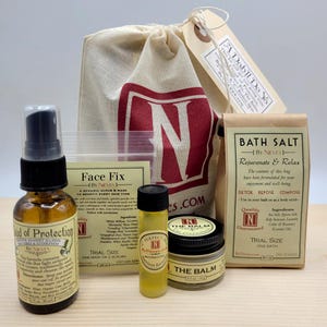 May include: A set of bath and body products in a natural canvas bag. The set includes a bottle of face fix, a bottle of cloud of protection, a small bottle of oil, a jar of balm, and a bag of bath salts. The products are all labeled with the brand name "By Nieves".