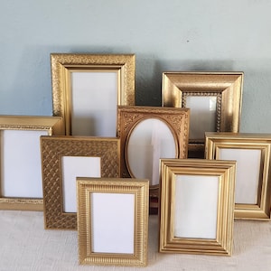 May include: A collection of eight gold-toned picture frames in various sizes and rectangular shapes, featuring diverse embossed and smooth border designs, each containing a blank white insert for displaying photographs or artwork.