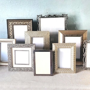 May include: A collection of nine assorted rectangular picture frames in various metallic finishes including gold, silver, and bronze. The frames feature diverse decorative textures, ornate borders, and intricate filigree details, each containing a blank white insert.