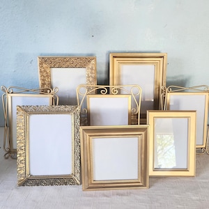 May include: A collection of eight gold-toned picture frames in various rectangular and square shapes. The frames feature diverse textures, including smooth, beaded, and ornate scrollwork patterns, each holding a blank white insert.