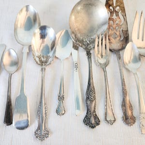 May include: A collection of assorted silver-toned metal flatware including various spoons, forks, and a serving utensil. The pieces feature diverse handle designs, ranging from plain polished surfaces to intricate floral and grape motifs.