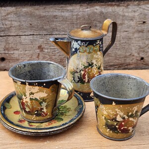 May include: Antique metal toy tea set with a decorative fruit design. The set includes a small teapot, two cups, and a saucer. The pieces have a distressed, aged appearance with gold, black, and yellow paint.