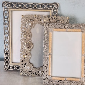 May include: Three rectangular picture frames with ornate metallic detailing. The back frame features a dark braided border with white beads. The middle frame has intricate silver scrollwork with embedded pearls and crystals. The front frame has a textured silver border with light tan panels.