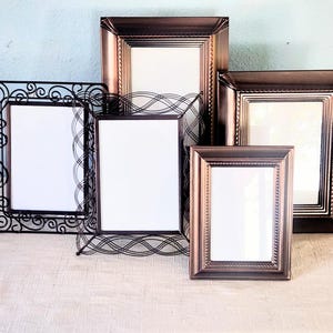 May include: A collection of five rectangular picture frames. Three frames are made of dark bronze-colored metal with beaded edges, while two frames feature intricate black wire designs with swirling and crisscross patterns.