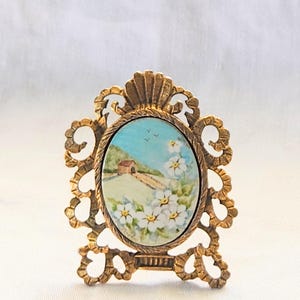 May include: Ornate gold-toned metal frame with an oval, hand-painted miniature landscape scene. The artwork depicts a bridge, a small building, and white flowers against a blue sky. The frame has decorative scrollwork and a shell-like top.