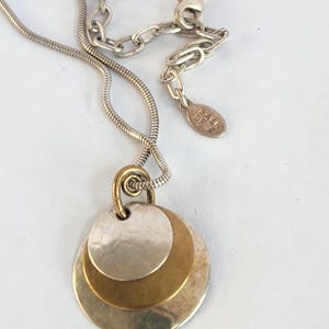 May include: Silver-toned necklace with a three-disc pendant. The pendant features two silver and one gold-toned disc, layered and connected by a small gold loop. The chain is a thin, snake-style design with a lobster clasp.