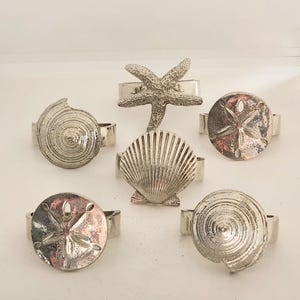 May include: A set of six silver-toned metal napkin rings featuring various marine life designs, including a starfish, a scallop shell, two spiral shells, and two sand dollars, each with a polished finish and a circular band attachment.