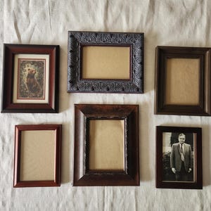 May include: A collection of six rectangular picture frames in various shades of brown and black. One frame contains a print of a bear, another holds a black and white photograph of a person in a suit, and four frames are empty with tan backings.