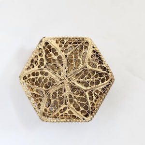 May include: A gold-colored, hexagonal box with intricate filigree detailing. The lid features a central floral design, and the edges are finely crafted. The box is set against a white background, highlighting its ornate design.
