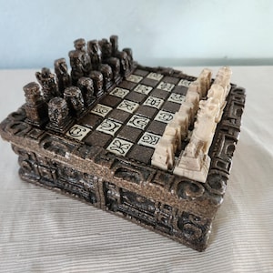 May include: A square chess set featuring a dark brown, textured base with carved geometric patterns. The board has alternating dark brown and off-white squares with etched symbols. One side has dark brown pieces, the other has off-white pieces.