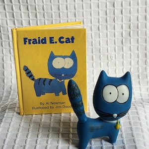 May include: A blue cat figurine with a striped tail and a yellow tag, next to a yellow book titled "Fraid E. Cat" by Al Newman, illustrated by Jim Dood. The book cover features a cartoon cat.