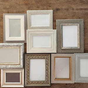 May include: A collection of various rectangular picture frames in different sizes and styles. The frames are in shades of white, gray, and gold, with ornate and simple designs. The frames are arranged on a wooden surface.