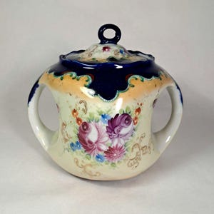 May include: A vintage porcelain sugar bowl with a lid. The bowl features a cream-colored base with hand-painted floral designs in shades of purple, pink, and blue. The rim is navy blue with a scalloped edge and a gold accent. The bowl has two handles.
