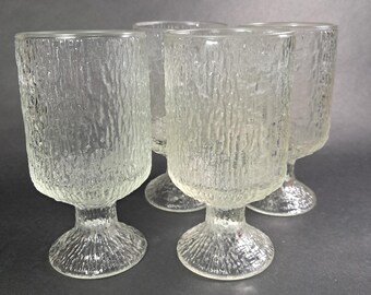 May include: Four clear, textured glass goblets with a stem and a wide, cylindrical bowl. The glass has a bark-like texture. The goblets are arranged closely together, with a dark gray background.