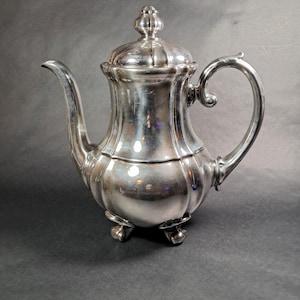 May include: A silver-colored teapot with a curved spout and handle. The teapot has a lid with a decorative finial and rests on three small feet. The design is ornate and reflective, suggesting a vintage or antique style.