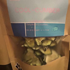 May include: A brown paper bag with a clear window showing dehydrated cucumber slices. The bag has a label that says "L&B's Treats & Sweets" and "Cool-Cumber Dehydrated Cucumbers Use as a topper or a treat".
