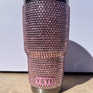 May include: A pink Yeti tumbler covered in rhinestones. The tumbler has a stainless steel base and a pink Yeti logo.