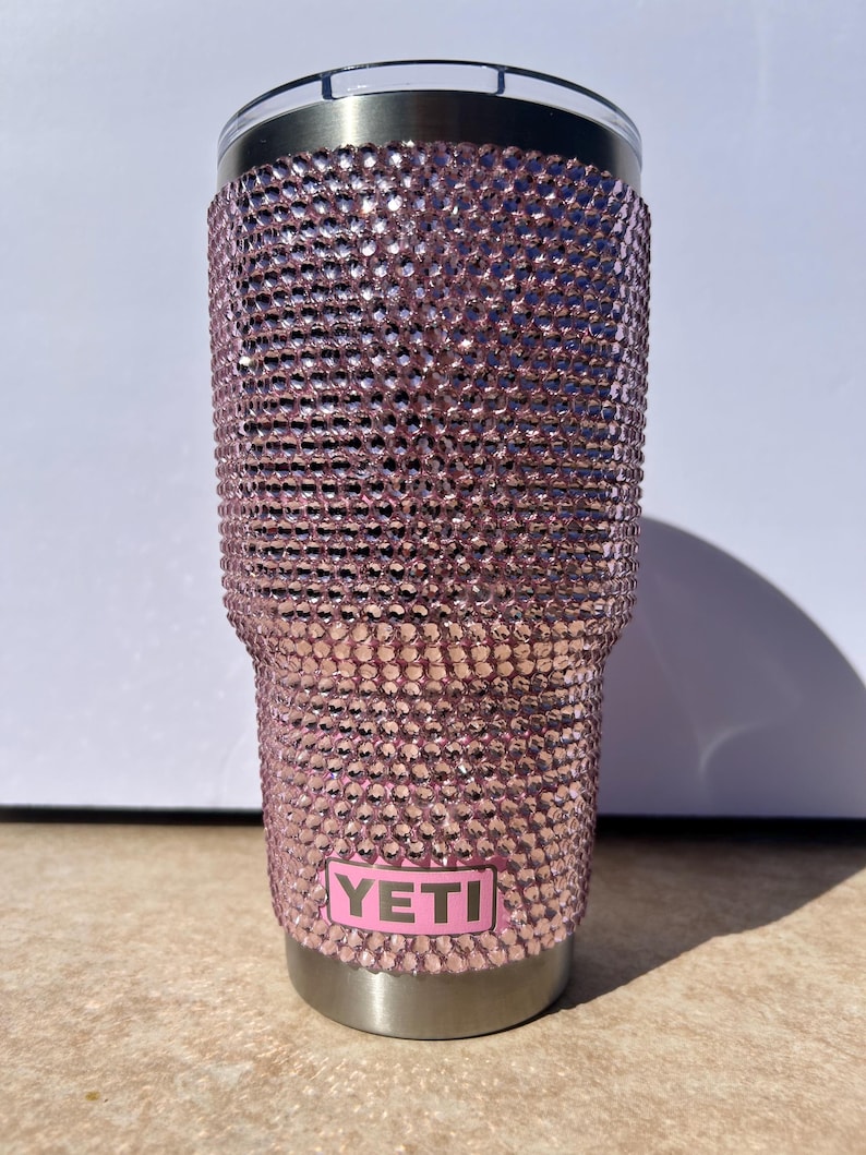 May include: A pink Yeti tumbler covered in rhinestones. The tumbler has a stainless steel base and a pink Yeti logo.