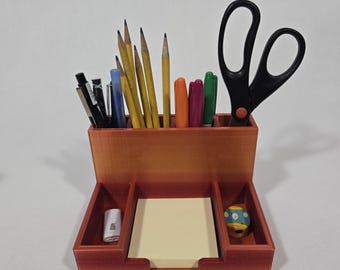 Può includere: Un organizzatore da scrivania in legno con più scomparti per penne, matite e altri articoli da ufficio. Ha un porta blocchi note integrato e un piccolo scomparto per un temperamatite. L'organizzatore è marrone con una tonalità rossastra.