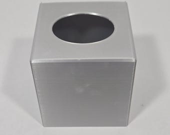 May include: A square, silver-coloured tissue box cover with a smooth, matte finish. The top surface features a centred, circular opening designed for dispensing tissues. The structure has clean, straight edges and a uniform, solid metallic appearance.