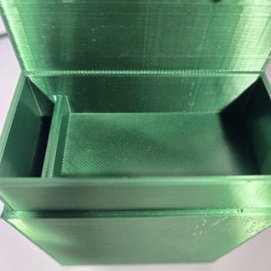 May include: A rectangular, emerald green storage box with a hinged lid and a single internal divider. The box features a visible 3D-printed texture with horizontal layer lines throughout the interior and exterior surfaces of the container.