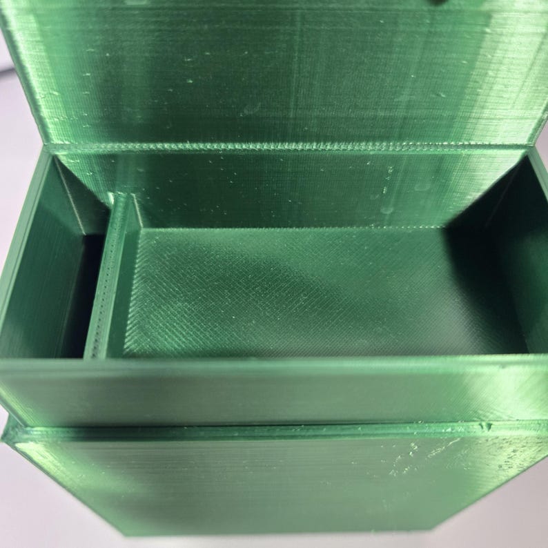 May include: A rectangular, emerald green storage box with a hinged lid and a single internal divider. The box features a visible 3D-printed texture with horizontal layer lines throughout the interior and exterior surfaces of the container.
