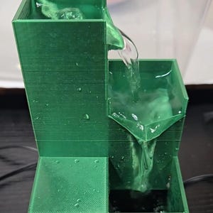 May include: A 3D-printed green tiered water fountain with a tall rectangular reservoir at the top, a middle basin, and a lower collection pool. Water flows from the top reservoir into the middle basin and then down into the bottom pool.