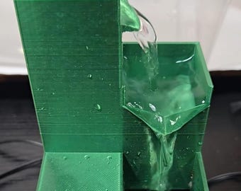 Può includere: Una fontana d'acqua a livelli stampata in 3D di colore verde, dotata di un alto serbatoio rettangolare nella parte superiore, un bacino intermedio e una vasca di raccolta inferiore. L'acqua scorre dal serbatoio superiore nel bacino centrale.