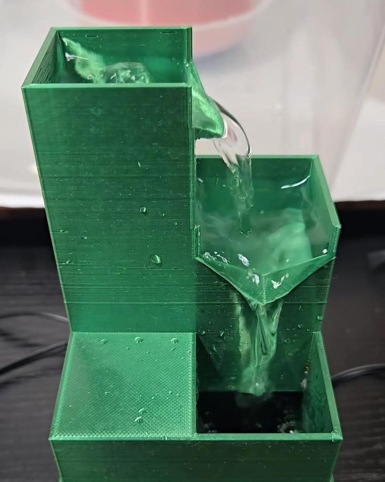 May include: A 3D-printed green tiered water fountain with a tall rectangular reservoir at the top, a middle basin, and a lower collection pool. Water flows from the top reservoir into the middle basin and then down into the bottom pool.