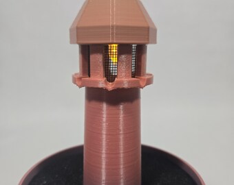 May include: A 3D printed lighthouse figurine in a muted mauve colour. The structure features a cylindrical base, a lantern room with a black mesh screen, and a pointed roof. A warm yellow light glows from within the lantern room, illuminating the mesh.