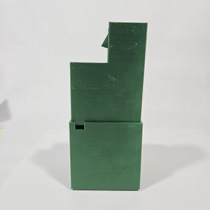 May include: A dark green, three-dimensional object with a stepped, rectangular structure. The item features a tall, narrow section on the right side and a shorter, wider base section on the left, displaying visible horizontal layer lines.