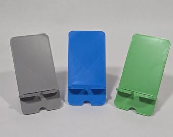 Può includere: Tre supporti rettangolari per telefono in grigio, blu e verde. Ogni supporto presenta un pannello posteriore piatto con una linea diagonale in rilievo, una piccola sporgenza orizzontale per il supporto del dispositivo e un intaglio semicircolare alla base.