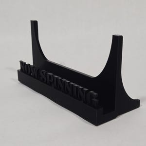 May include: A black vinyl record display stand with the words "NOW SPINNING" in raised lettering. The stand has a rectangular base and two curved supports. The stand is designed to hold a single record.